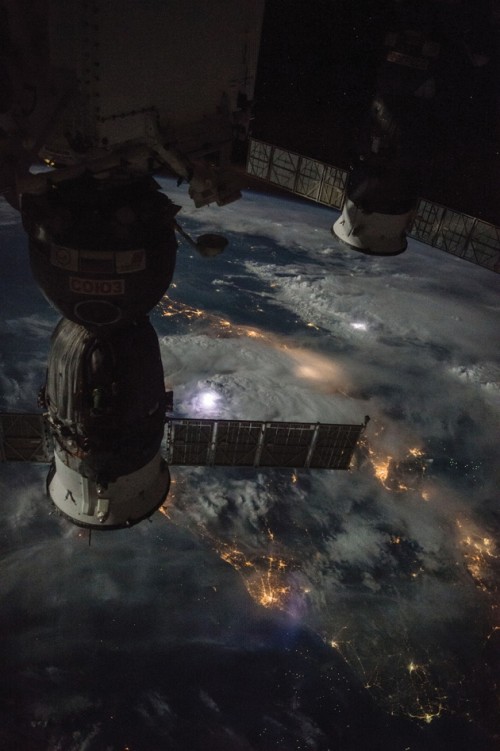 International Space Station, lightning flashes illuminate the clouds, as human activity is revealed by clusters of lights.