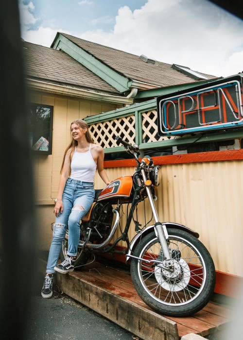 Woman in white cami top leaning on orange motorcycle